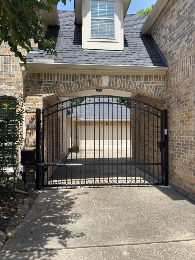 Arched spear-top iron gate fitted under a brick porte-cochere with a LiftMaster arm operator, installed in Cypress, TX