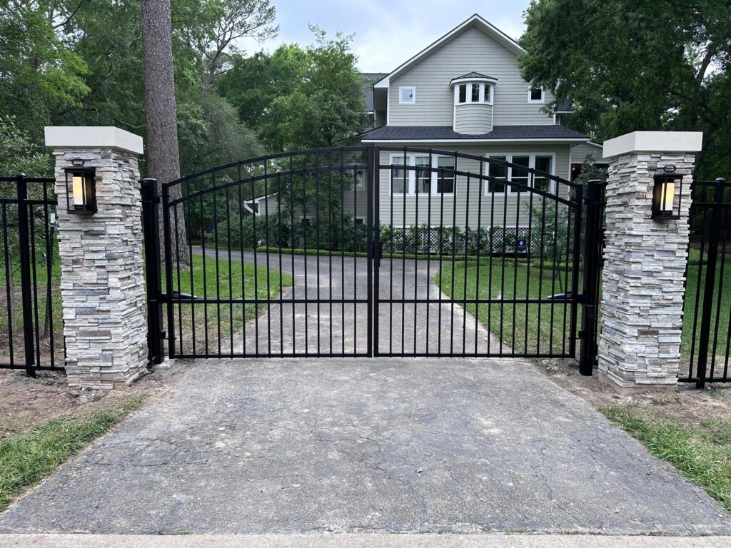 Arched iron double driveway gate between stacked stone columns with lanterns, installed in Houston, TX