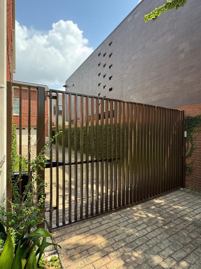 Bronze vertical-slat metal driveway gate on a brick paver drive, installed in Katy, TX