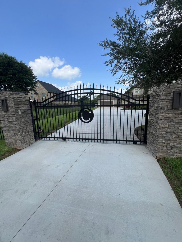 Arched spear-top wrought iron driveway gate with a custom-cut monogram between stacked stone columns, installed in Magnolia, TX
