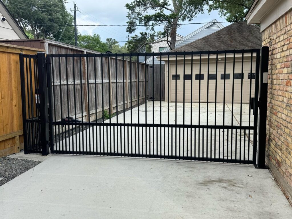 Black iron driveway gate shown open beside a wood fence and detached garage, installed in Pearland, TX