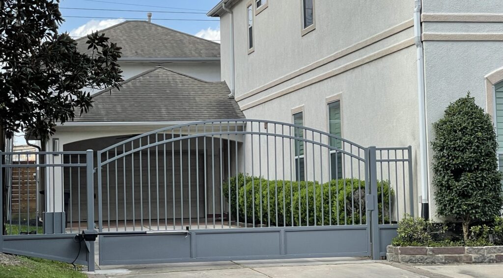 Grey painted steel driveway gate with an arched top and matching fence at a stucco home in Bellaire, TX